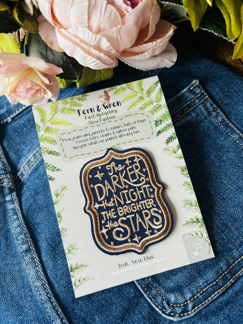 May include: A navy blue embroidered patch with a decorative border and gold lettering that reads, The DARKER the NIGHT the BRIGHTER the STARS. The patch is mounted on a white card featuring green fern illustrations and text about upcycling.