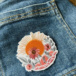 Puede incluir: Parche bordado con un sol naranja vibrante con l&iacute;neas radiantes, sobre un fondo de flores y follaje rosas y grises. El parche est&aacute; sobre un fondo de mezclilla azul, mostrando el dise&ntilde;o y los colores del parche.