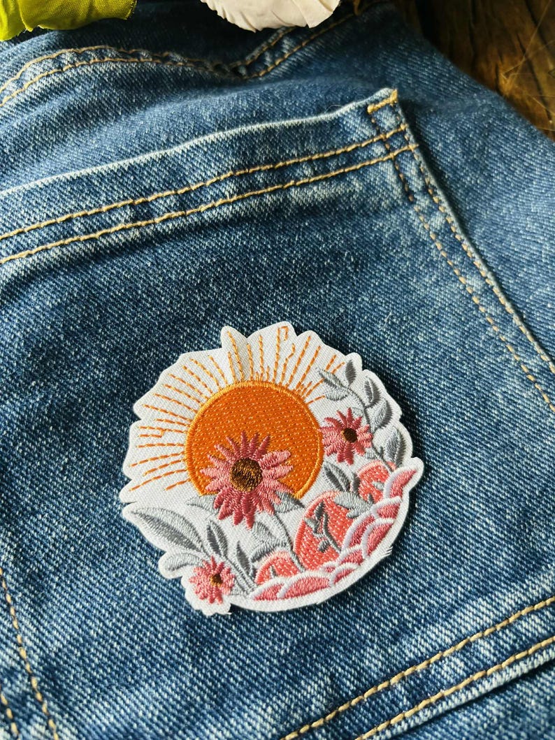 Puede incluir: Parche bordado con un sol naranja vibrante con l&iacute;neas radiantes, sobre un fondo de flores y follaje rosas y grises. El parche est&aacute; sobre un fondo de mezclilla azul, mostrando el dise&ntilde;o y los colores del parche.