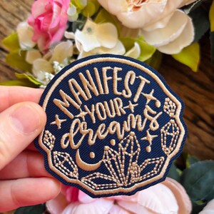 May include: A circular navy blue embroidered patch featuring the gold-colored text "MANIFEST YOUR dreams" in a mix of block and script fonts. The design includes gold-stitched crystal clusters, a crescent moon, and small star accents.
