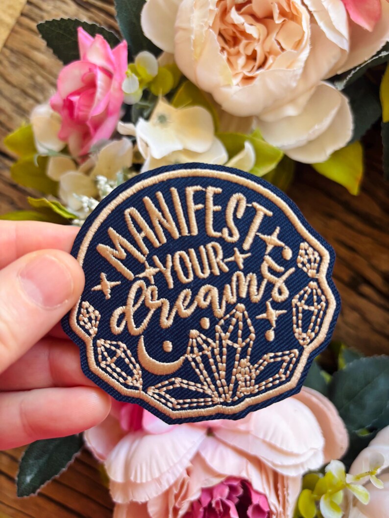 May include: A circular navy blue embroidered patch featuring the gold-colored text "MANIFEST YOUR dreams" in a mix of block and script fonts. The design includes gold-stitched crystal clusters, a crescent moon, and small star accents.