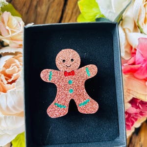 May include: A glittery, reddish-brown gingerbread man brooch with a red bow tie and green accents, presented in a black box. The brooch is surrounded by colorful flowers, suggesting a festive or holiday theme.