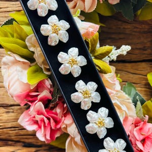 May include: Six white flower-shaped earrings with gold centers are displayed in a black box. The earrings have a pearl-like appearance. The background features a floral arrangement with pink and white flowers and green leaves.