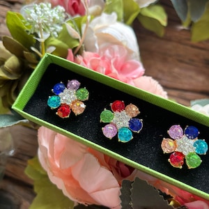 May include: Three colorful flower-shaped brooches with rhinestones in a green and black box. The brooches are gold-toned and have a rainbow of colors.