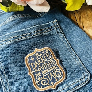 May include: A rectangular embroidered patch with a scalloped border attached to a blue denim pocket. The patch features a dark blue background with gold-colored text that reads, The DARKER the NIGHT the BRIGHTER the STARS, accented by small stars.
