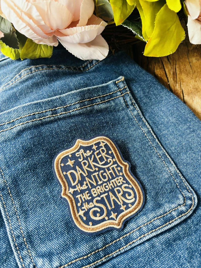 May include: A rectangular embroidered patch with a scalloped border attached to a blue denim pocket. The patch features a dark blue background with gold-colored text that reads, The DARKER the NIGHT the BRIGHTER the STARS, accented by small stars.