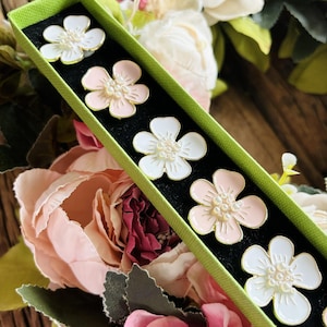 May include: A set of six decorative flower-shaped buttons in a green box. The buttons are white and pink with pearl-like accents.