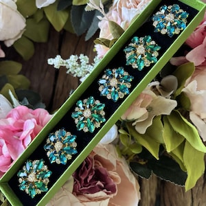 May include: A set of six jeweled brooches in a green box. Each brooch features a floral design with teal and emerald green crystals and a central cluster of clear crystals. The brooches are set against a black background within a green box.