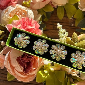 May include: Four iridescent flower-shaped brooches with clear rhinestones in a green velvet box. The box is surrounded by pink and white flowers.