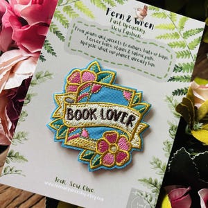 May include: An embroidered patch featuring a light blue background with pink flowers and green leaves. A central white banner with a gold border displays the text "BOOK LOVER" in dark brown letters. The patch is attached to a branded display card.