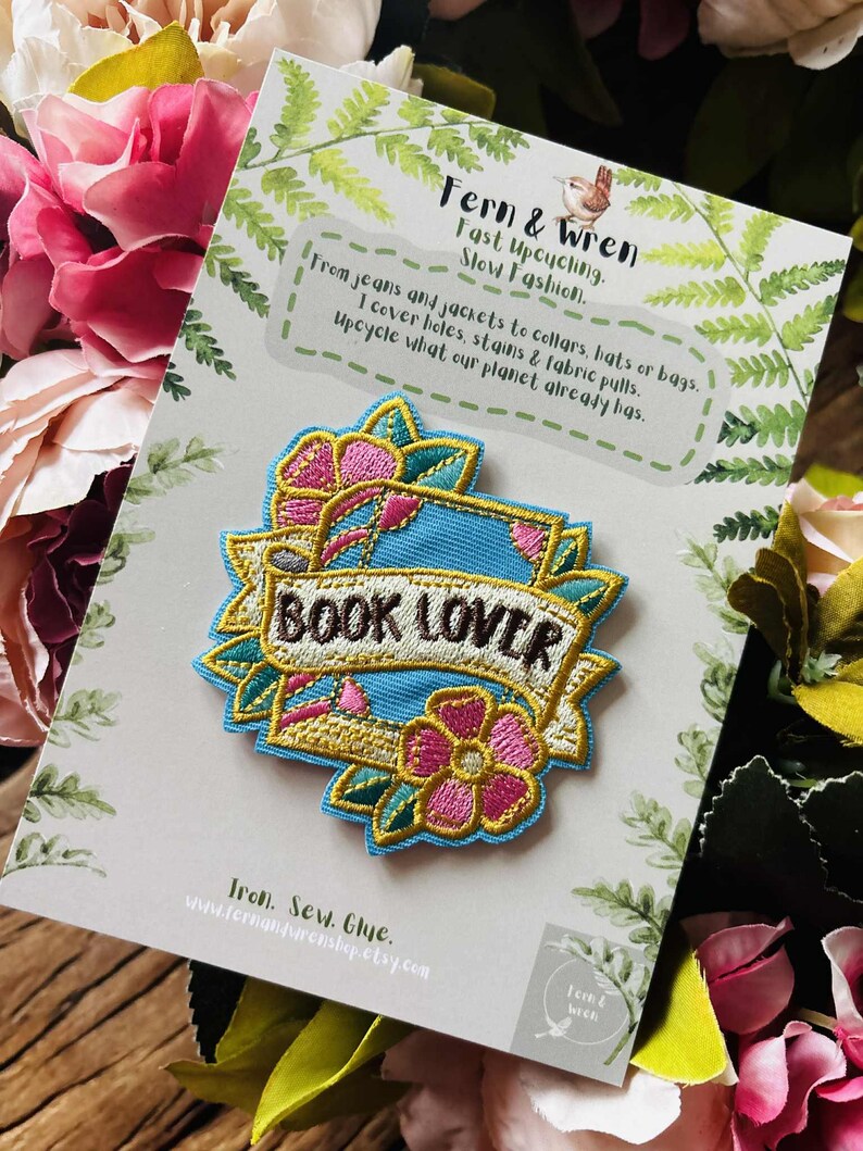 May include: An embroidered patch featuring a light blue background with pink flowers and green leaves. A central white banner with a gold border displays the text "BOOK LOVER" in dark brown letters. The patch is attached to a branded display card.