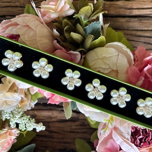 May include: Six flower-shaped earrings with pearl petals and a central rhinestone are displayed in a green box with a black interior. The earrings are set against a backdrop of pink and white flowers.