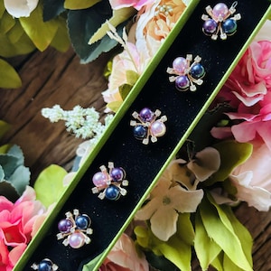 May include: A set of six decorative brooches in a green box. Each brooch features a cluster of purple, pink, and black pearls with gold accents.