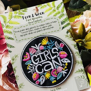 May include: A circular black embroidered patch featuring the white text GIRLS CAN surrounded by colorful floral motifs in pink, yellow, and light blue. The patch is attached to a white display card with green fern illustrations and text.