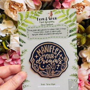 May include: A circular navy blue embroidered patch featuring the gold text "MANIFEST YOUR dreams" surrounded by crystal and moon motifs. The patch is attached to a light green card with fern illustrations and text about upcycling.