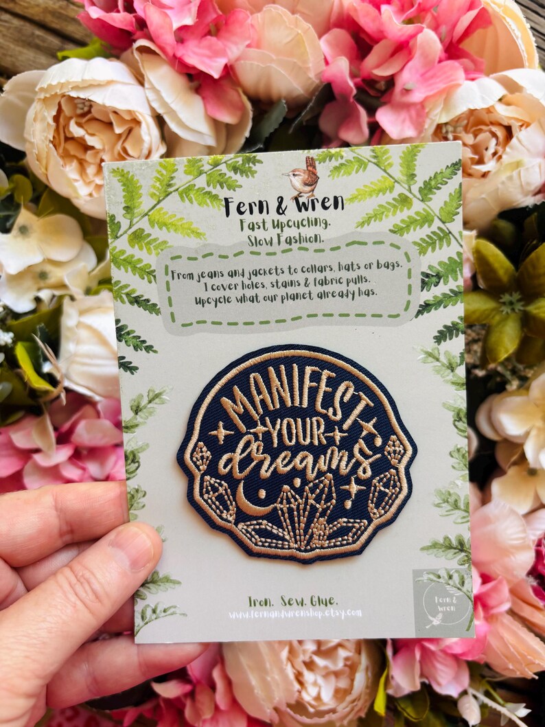 May include: A circular navy blue embroidered patch featuring the gold text "MANIFEST YOUR dreams" surrounded by crystal and moon motifs. The patch is attached to a light green card with fern illustrations and text about upcycling.