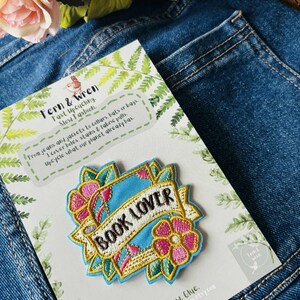 May include: An embroidered patch featuring the text BOOK LOVER in black letters on a light blue banner, surrounded by pink and yellow flowers and green leaves. The patch is attached to a white display card with green fern illustrations and text.