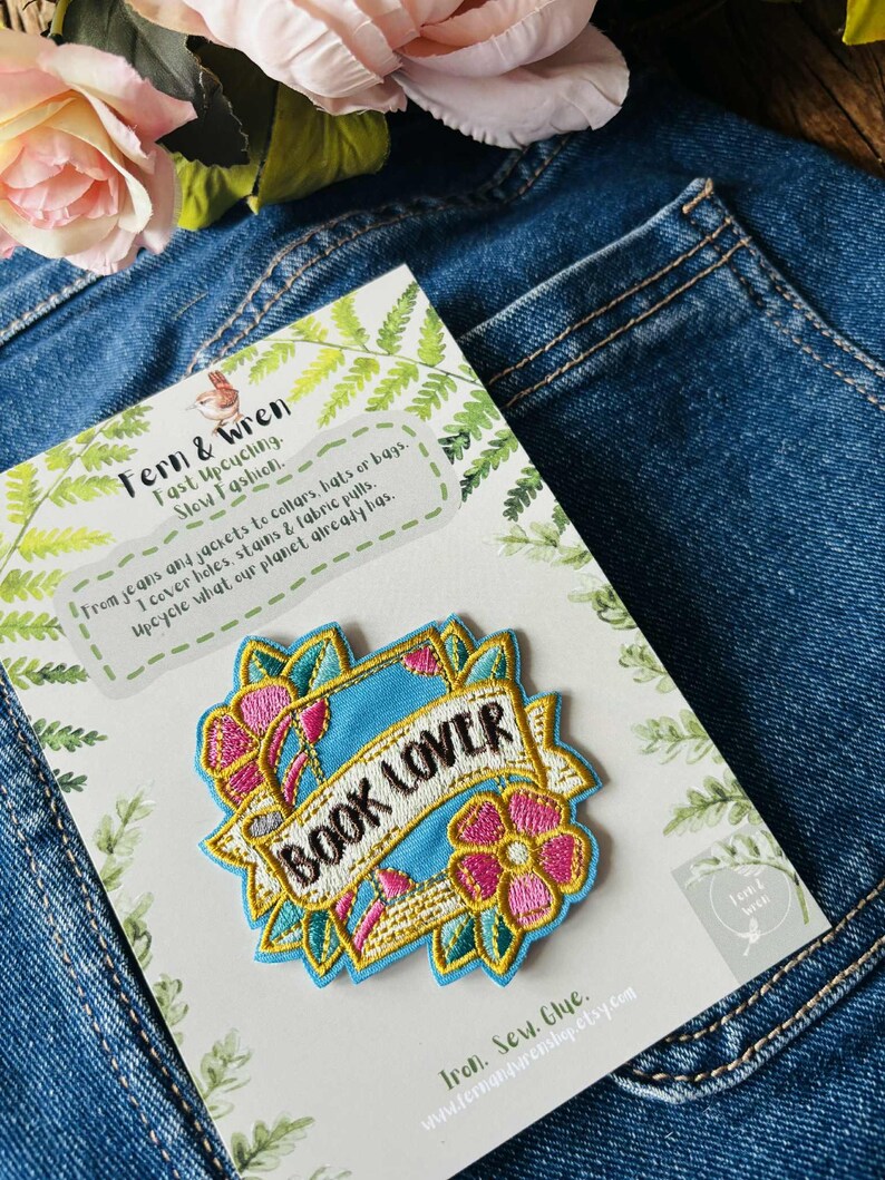May include: An embroidered patch featuring the text BOOK LOVER in black letters on a light blue banner, surrounded by pink and yellow flowers and green leaves. The patch is attached to a white display card with green fern illustrations and text.