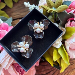 May include: A pair of flower-shaped earrings in a black box. Each earring features a dark brown, petal-like design with a cluster of white pearls and gold-colored accents. The earrings are set against a backdrop of colorful flowers.