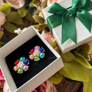 May include: A pair of flower-shaped stud earrings with colorful gemstones, presented in a white gift box with a green ribbon. The earrings feature red, orange, yellow, green, blue, and purple stones. The background includes flowers.