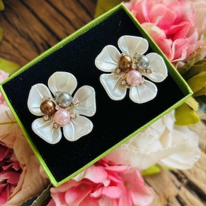 May include: A pair of flower-shaped earrings with white petals and pearl centers in shades of brown, gray, and pink. The earrings are presented in a green-trimmed black box, surrounded by pink and white flowers.