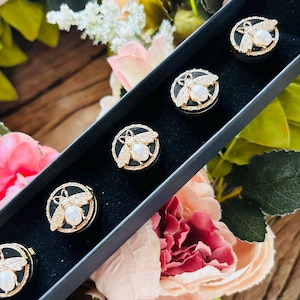 May include: A set of six gold-toned bee-shaped buttons with pearl-like centers, displayed in a black box. The bees have detailed wings and are set against a black background. The box is surrounded by pink and green floral arrangements.