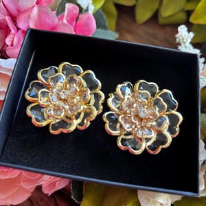 May include: A pair of gold-toned flower-shaped earrings with black and clear crystal accents, presented in a black box. The earrings have a layered petal design and a central cluster of clear crystals. The background features pink and white flowers.