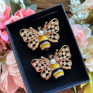 May include: Two gold-toned bee brooches with rhinestone accents and pearl-like beads. The bees have yellow and black striped bodies. The brooches are displayed in a black box, surrounded by pink and green floral decorations.