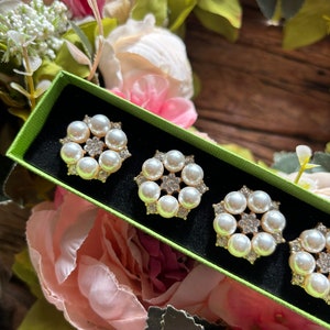 May include: Four gold-toned brooches with white pearl accents in a green box. The brooches are arranged in a row, with the pearls forming a circular pattern.