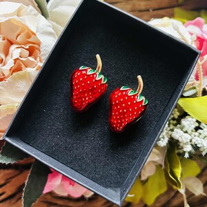 May include: A pair of red strawberry-shaped earrings with green leaves and gold stems, presented in a black gift box. The earrings have a glossy finish and are set against a backdrop of flowers.