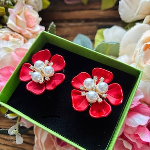 May include: A pair of red flower-shaped earrings with pearl and rhinestone accents, presented in a green box with a black interior. The earrings are set against a backdrop of soft-focus flowers.