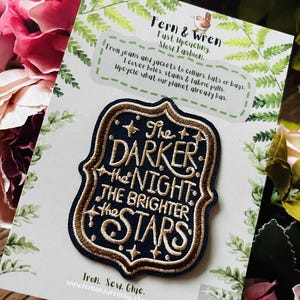 Könnte beinhalten: Gestickter Aufnäher mit dem Text "The Darker the Night the Brighter the Stars" in Gold und Weiß auf marineblauem Hintergrund. Der Aufnäher hat eine dekorative Schildform und ist von einem goldenen Rand umgeben. Der Aufnäher befindet sich auf einer weißen Karte mit Text zum Thema Upcycling.