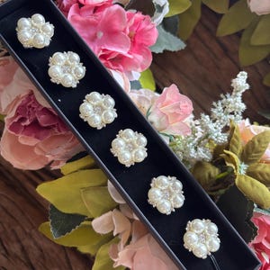 May include: A set of six pearl and rhinestone hair pins in a black box. Each pin features a cluster of faux pearls surrounding a central rhinestone. The pins are arranged in a row, with a single pin at the end.