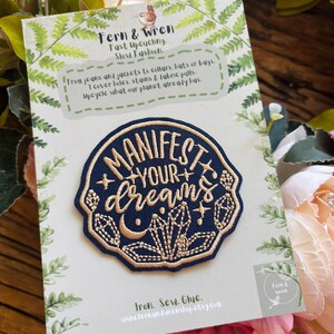 May include: A circular embroidered patch featuring a dark blue background with gold stitching that reads "MANIFEST YOUR dreams" surrounded by crystal and moon motifs. The patch is attached to a white card with green fern leaf illustrations.