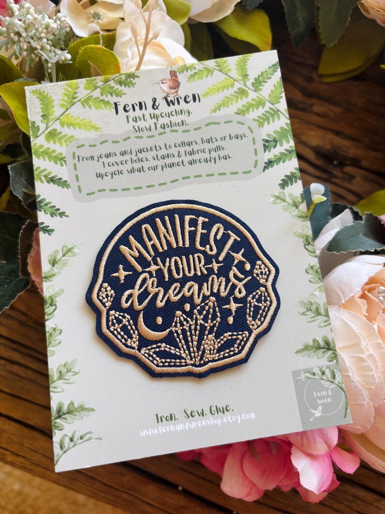 May include: A circular embroidered patch featuring a dark blue background with gold stitching that reads "MANIFEST YOUR dreams" surrounded by crystal and moon motifs. The patch is attached to a white card with green fern leaf illustrations.