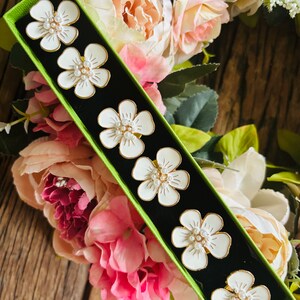 May include: A green box with a white background contains six white ceramic flower-shaped napkin rings with gold trim and pearl accents.