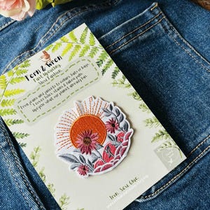 Puede incluir: Parche bordado con un dise&ntilde;o de sol y flores. El parche tiene un fondo blanco con detalles en naranja, rosa y gris. El parche est&aacute; sobre una tarjeta con el texto "Fern & Wren" y "Fast Upcycling. Slow Fashion."