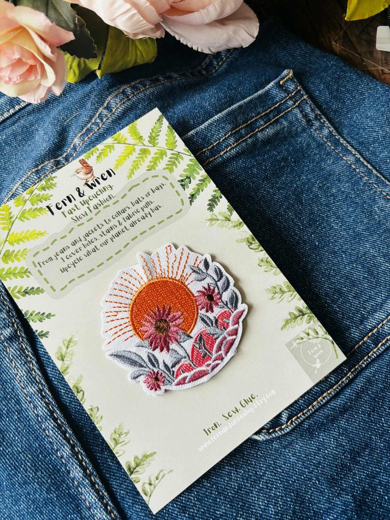 Puede incluir: Parche bordado con un dise&ntilde;o de sol y flores. El parche tiene un fondo blanco con detalles en naranja, rosa y gris. El parche est&aacute; sobre una tarjeta con el texto "Fern & Wren" y "Fast Upcycling. Slow Fashion."