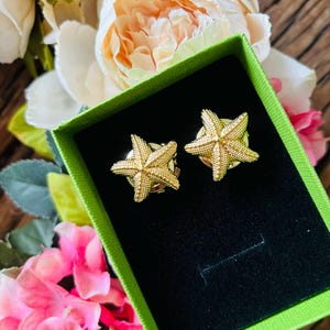 May include: A pair of gold-colored starfish earrings displayed in a green box with a black interior. The earrings are detailed with textured surfaces, and the background features pink and white flowers.