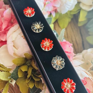 May include: A set of six flower-shaped buttons in a black box. The buttons alternate between red and black petals with gold centers. The box is displayed against a backdrop of pink and white flowers.