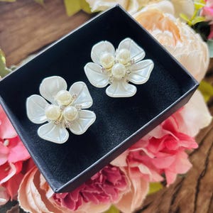 May include: Two white flower-shaped earrings with pearl centers and rhinestone accents are displayed in a black box. The earrings are set against a backdrop of pink and cream-colored flowers.