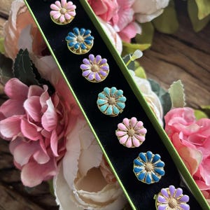 May include: A set of eight colorful daisy-shaped buttons in a green box. The buttons are arranged in a row and feature different colors, including pink, blue, purple, and teal.
