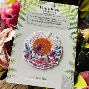 Puede incluir: Parche bordado con un dise&ntilde;o de puesta de sol, que incluye un sol naranja, flores rosas y hojas verdes. El parche est&aacute; sobre una tarjeta con el texto "Fern & Wren, Fast Upcycling, Slow Fashion". El parche se puede planchar, coser o pegar.