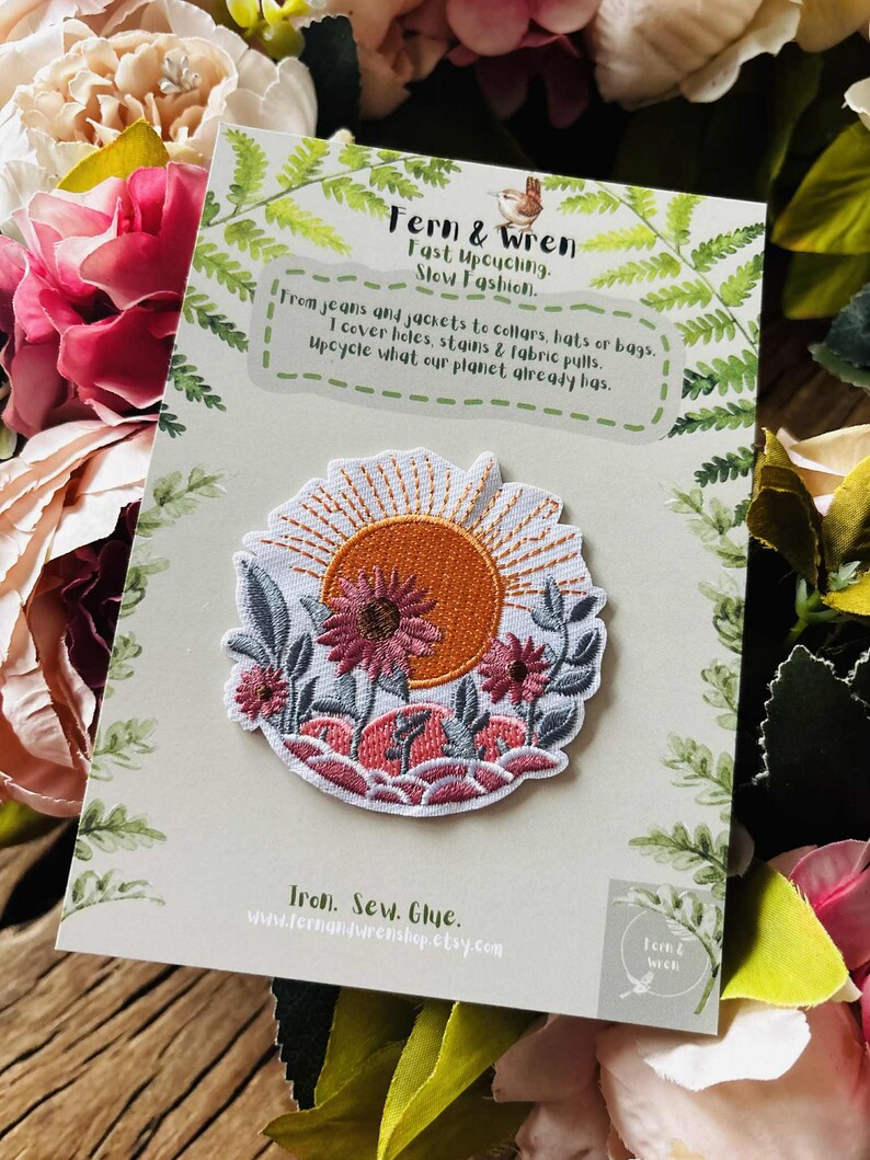 Puede incluir: Parche bordado con un dise&ntilde;o de puesta de sol, que incluye un sol naranja, flores rosas y hojas verdes. El parche est&aacute; sobre una tarjeta con el texto "Fern & Wren, Fast Upcycling, Slow Fashion". El parche se puede planchar, coser o pegar.