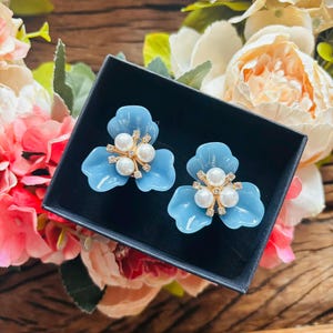 May include: A pair of light blue flower-shaped earrings with pearl and rhinestone accents, presented in a black box. The earrings are set against a backdrop of colorful flowers, including pink and white peonies.