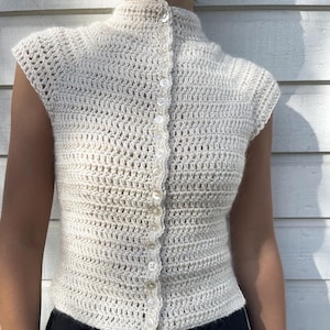 Puede incluir: Un top de crochet sin mangas de color crema, con cuello alto y cierre de botones en la parte delantera. El top está hecho de un hilo texturizado de tejido abierto y presenta pequeños botones redondos. La prenda se lleva con pantalones negros.