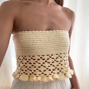 May include: Cream-colored crochet tube top. The top features a solid, textured band at the top, a decorative openwork pattern in the middle, and a ruffled edge at the bottom. The garment is handmade and has a bohemian aesthetic.