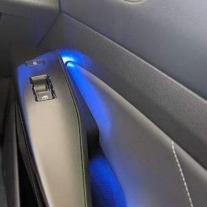 May include: A close-up of a car door interior with a blue ambient light illuminating the door handle and the window controls. The door panel is gray and has a black leather-like texture.
