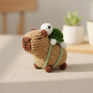 May include: A crocheted capybara toy with a green turtle riding on its back. The capybara is a light brown colour with darker brown details, and the turtle has a white head and green shell. The two are connected with a green strap. A whimsical amigurumi.