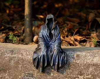 Könnte beinhalten: Dunkelgraue Statue einer Kapuzenfigur, die auf einer Betonleiste sitzt. Die Figur ist in fließende Roben gehüllt, mit einer gesichtslosen Kapuze. Die Statue hat ein verwittertes Aussehen, was darauf hindeutet, dass sie aus Stein oder Metall besteht. Der Hintergrund ist verschwommen, mit Andeutungen von Laub.